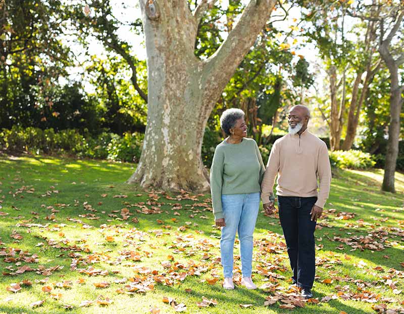 A senior Black couple, holding hands, smiles at each other as they walk through a sunny park filled with green grass and fallen autumn leaves.