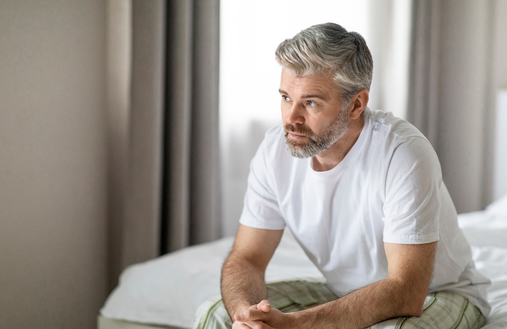 Pensive thoughtful unhappy grey-haired bearded mature man wearing pajamas sitting on bed at home feeling lonely.